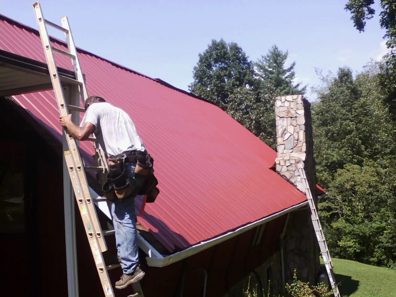 Roofer on a ladder beside a red metal roof and stone chimney — Soffit & Fascia Repair by Roofing Brothers in Irondale